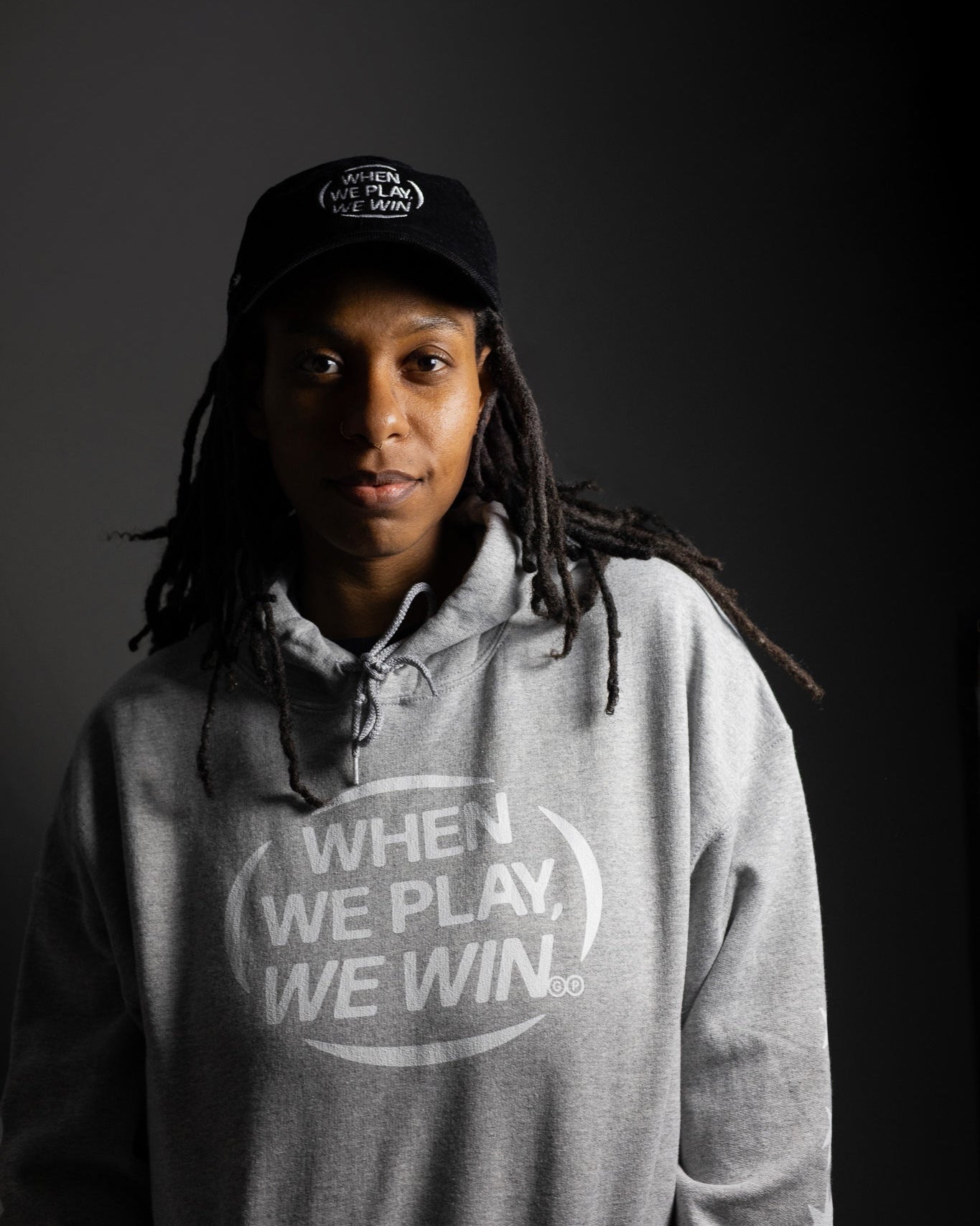Person wearing a gray hoodie with 'When We Play, We Win' text and a black cap on a dark background
