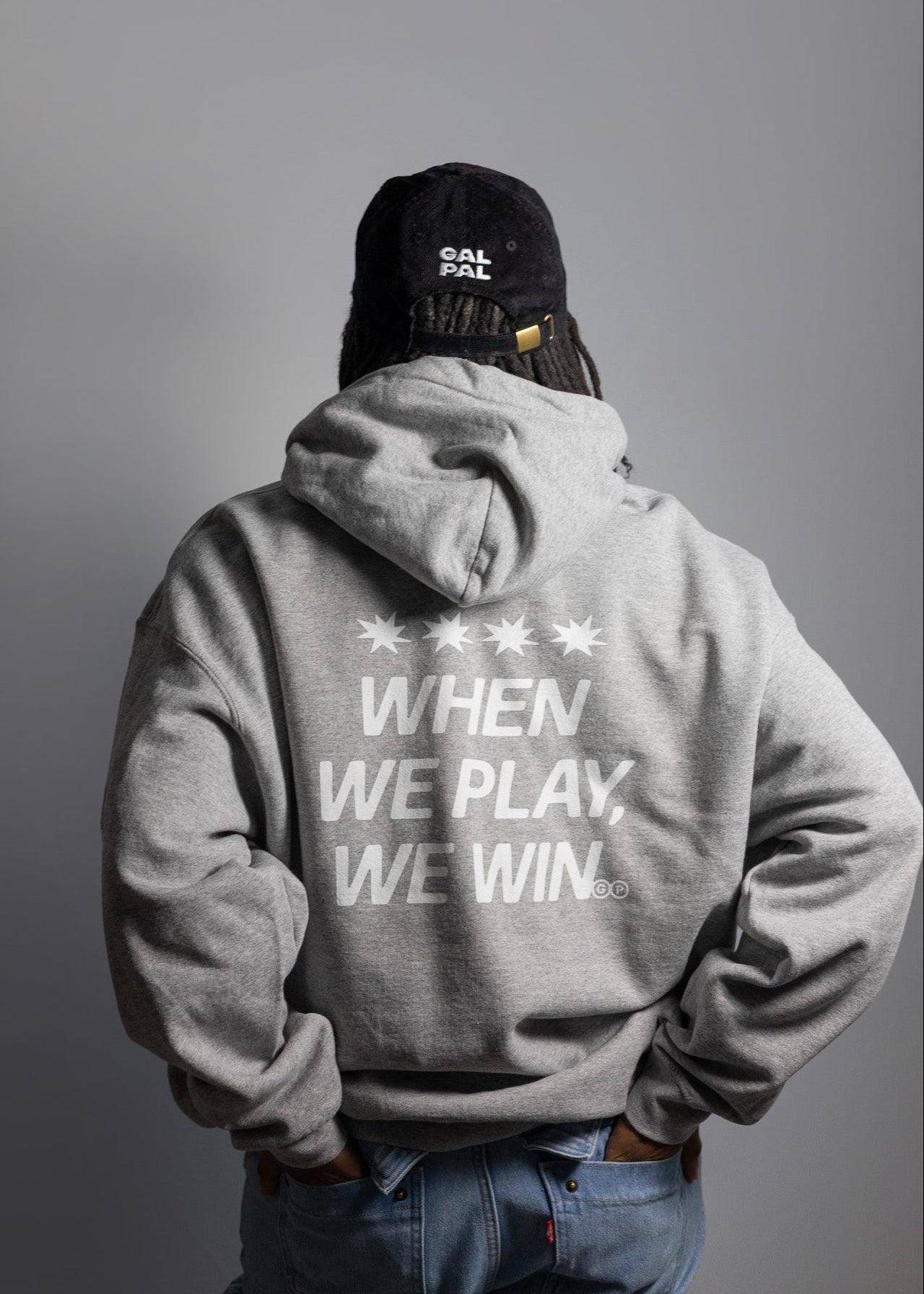 Person wearing a soft gray hoodie with "When We Play We Win" text and a black cap on a plain background