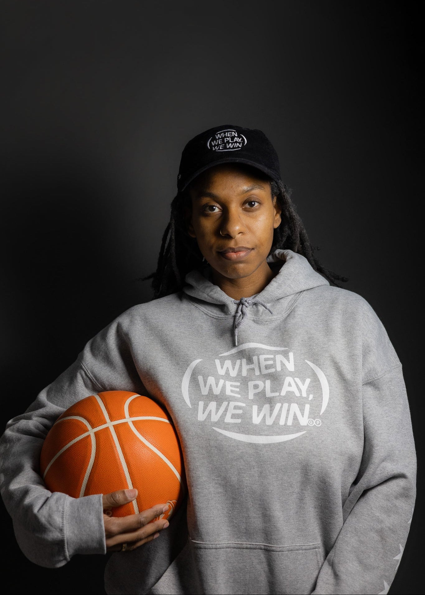Person wearing a gray hoodie with a basketball and cap on a dark background